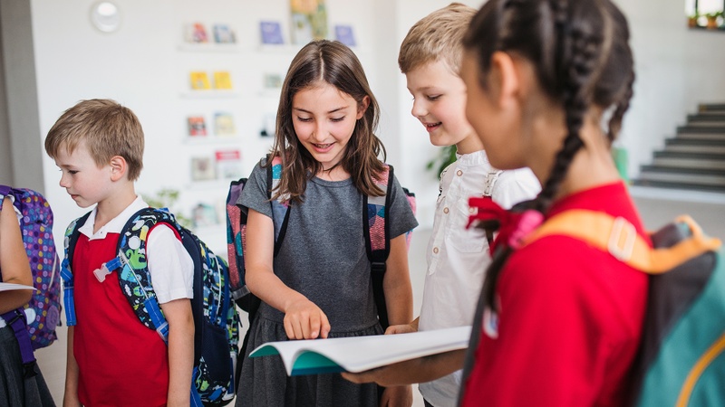 Schulkinder stehen im Flur des Schulhauses, ein Mädchen zeigt einem Jungen etwas in einem Heft